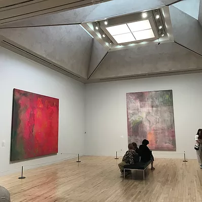 a zoomed out photo of one corner of the exhibition with people sat on a bench to put the scale of the paintings into perspective, they look metres high