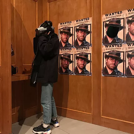 Zarina stands in a wooden booth wearing a VR headset with images of Arthur Morgan on a Wanted sign behind her - Arthur is from Red Dead Redemption 2