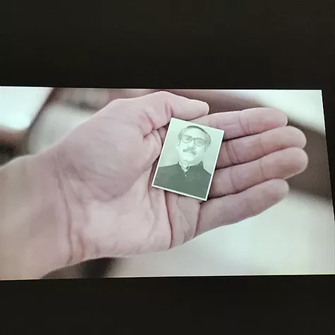 a hand holds a tiny passport picture of a man with a moustache and glasses