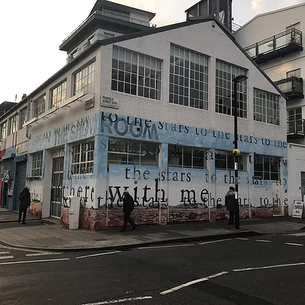 the outside of the gallery, which looks like a warehouse with big windows, has a brick wall painted on the bottom and a blue sky up to halfway with words faint and repeated across the image like &lsquo;with me, the stars, to the stars, there with me&rsquo;