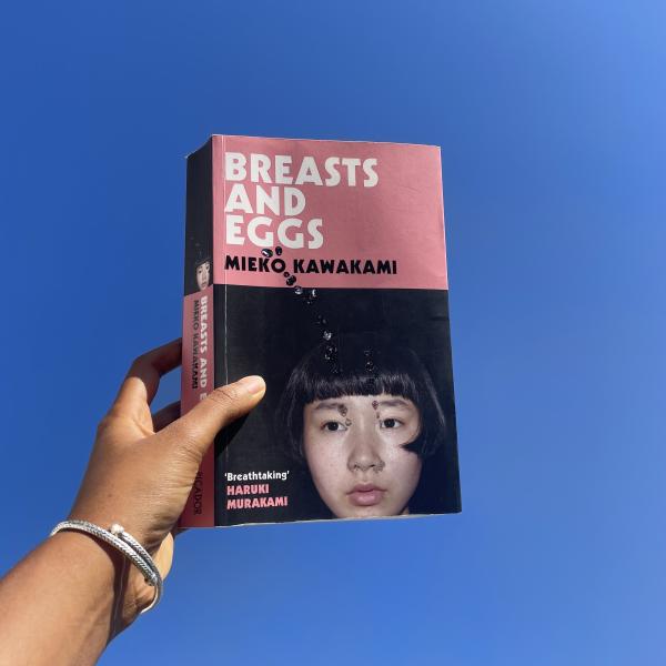 zm’s hand holding a copy of breasts and eggs up to the sky. The sky is blue and cloudless, and the book cover is pink at the top, and there is a picture of a girl with a blunt fringe on the bottom half