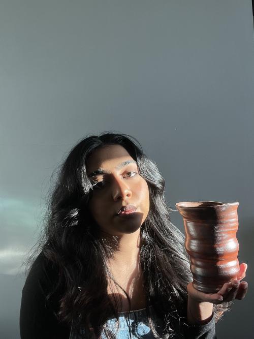 francesca is half lit holding a brown vase that rises up in undulating shapes getting wider and then narrower and wider, but in an uneven pattern with an uneven edge