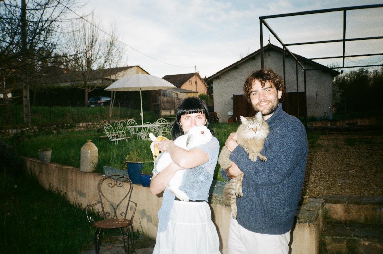Tahney and Reece holding a rabbit called Enzo and a cat called Maahs in front of the Mill, where you can see garden furniture and the workshop in the background