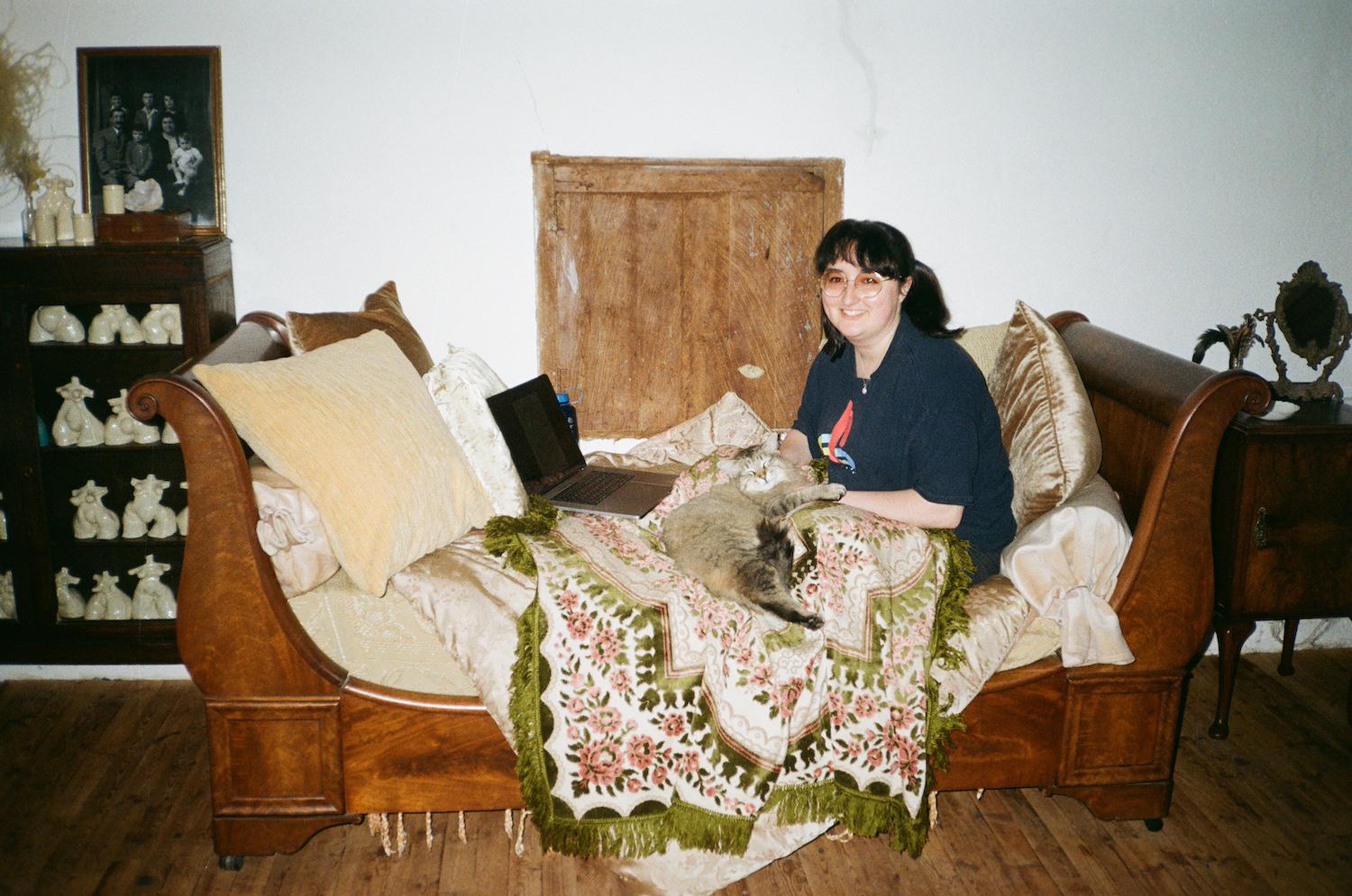 Gabrielle smiling on an antique day bed under blankets with a laptop and a beautiful cat called Maahs on her lap, who looks like a Norwegian forest cat or a Siberian cat
