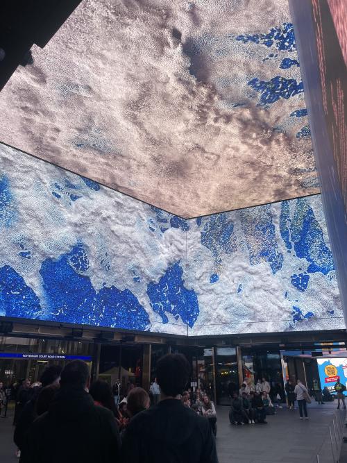 blue cyanotype flags hanging in aa gallery showing ursula k le guin's maps, plus zarina with her arms folded to one side of the picture